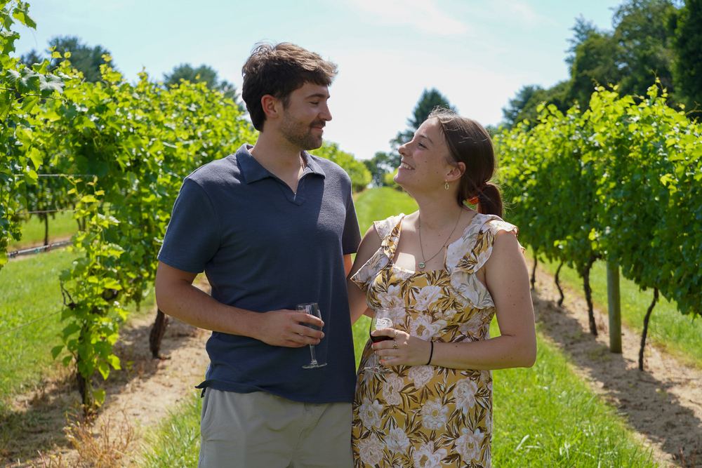 Couple smiling at each other with wine glasses in a Maryland vineyard, engagement photography by Campbell Shepard