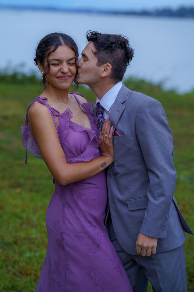 Person in a gray suit kissing the cheek of a person in a purple dress beside water