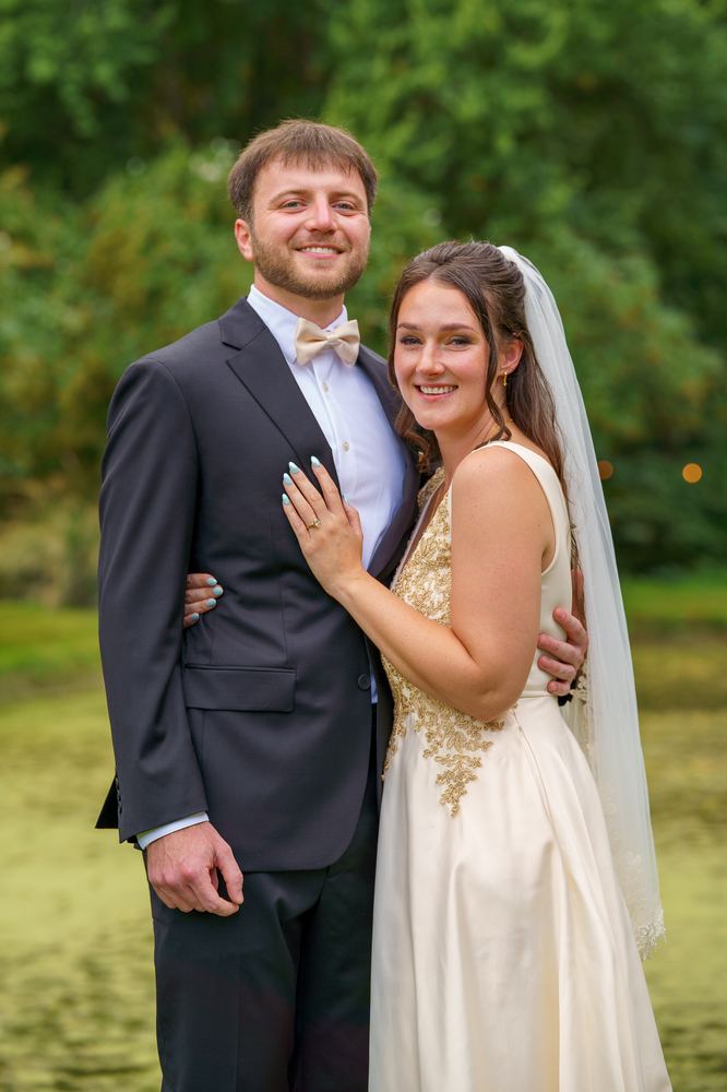 Two people in wedding attire standing together in front of trees